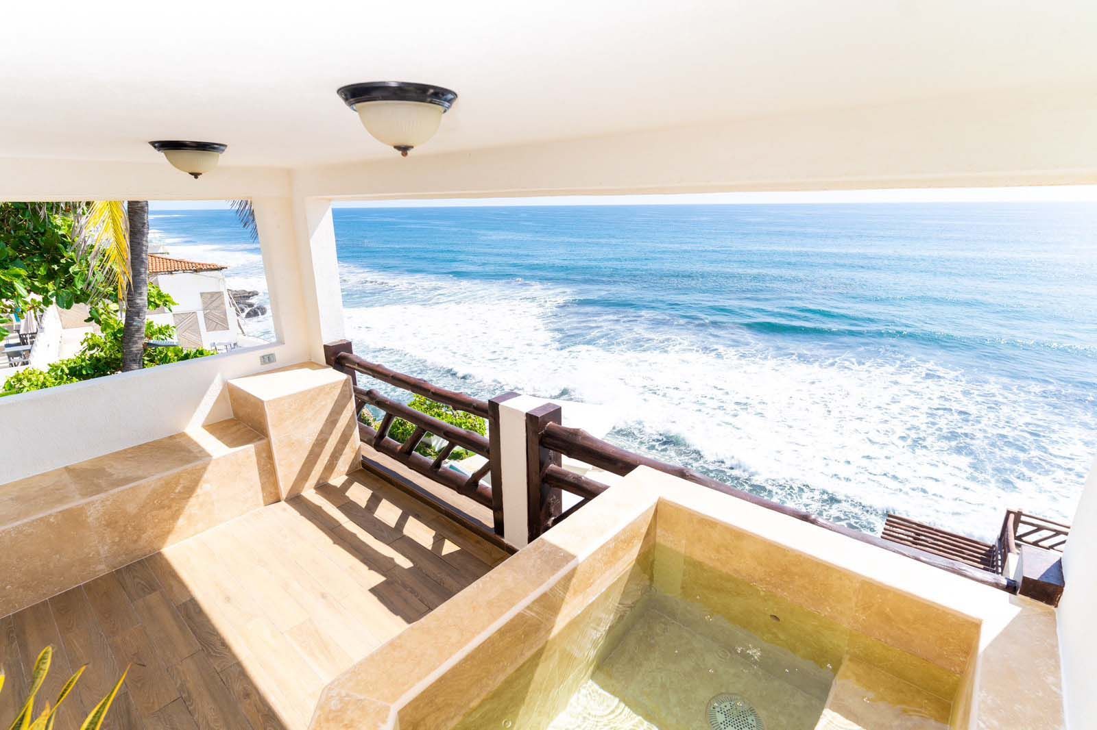 A stone-tiled balcony overlooking the ocean, featuring a built-in tub, wooden railings, and two ceiling dome lights.