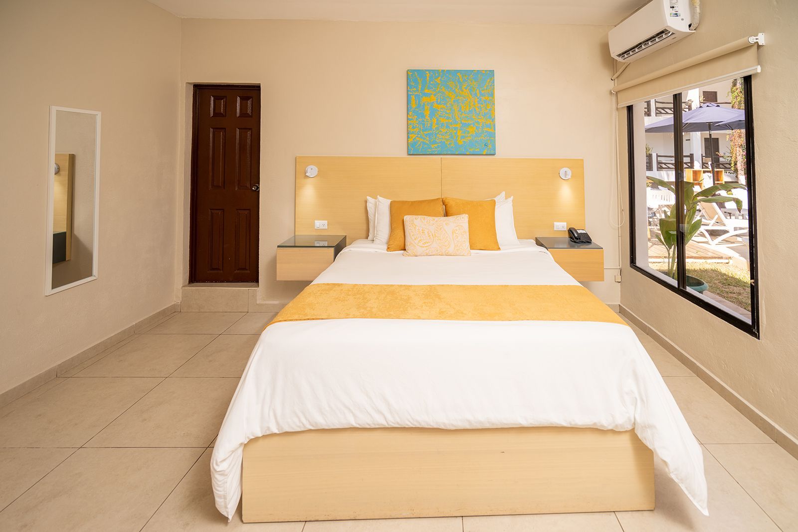 A clean hotel room with a queen-sized bed, a yellow runner, a matching headboard, a window, and a dark wooden door.