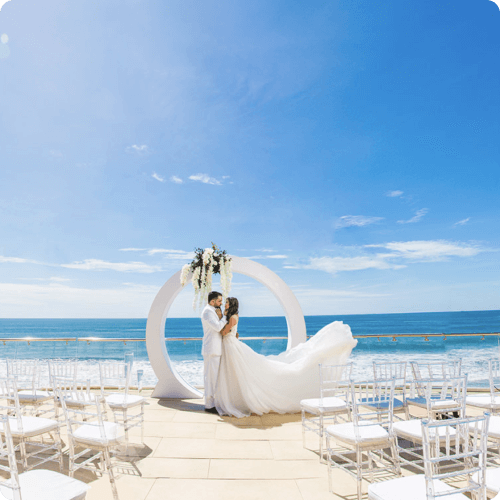 A wedding couple stands under a circular arch on a beach terrace with rows of white chairs facing the ocean.
