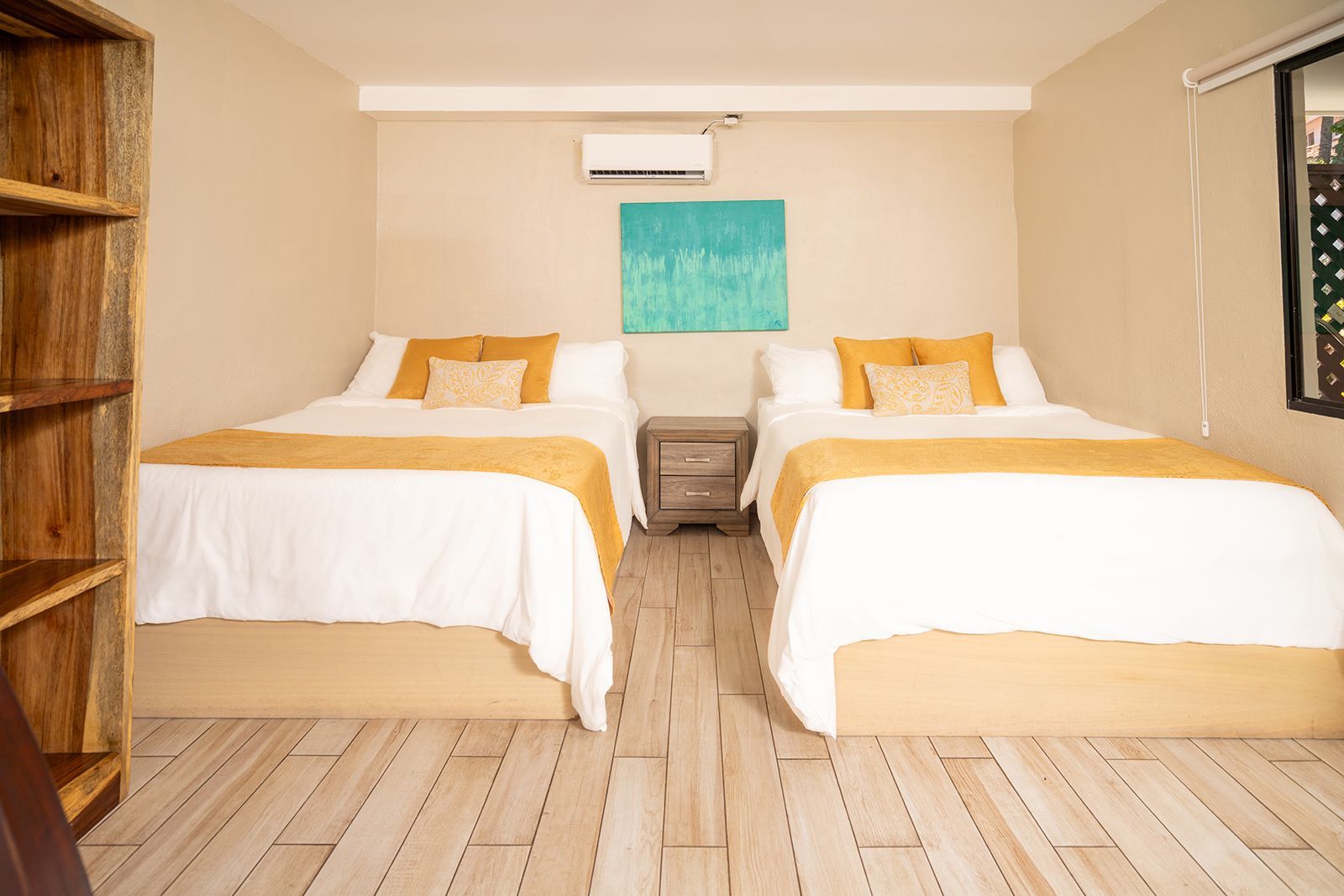 Two beds with white linens and mustard-yellow accents sit in a room with a light wooden floor and a small nightstand.