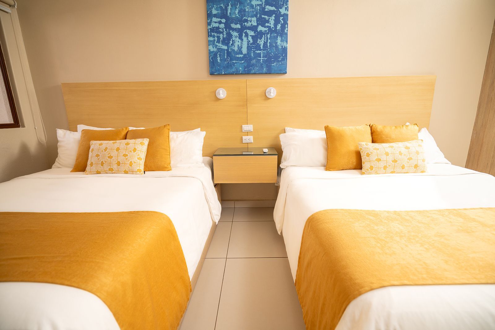 A hotel room featuring two beds with white linens, gold runners, patterned accent pillows, and a central nightstand.