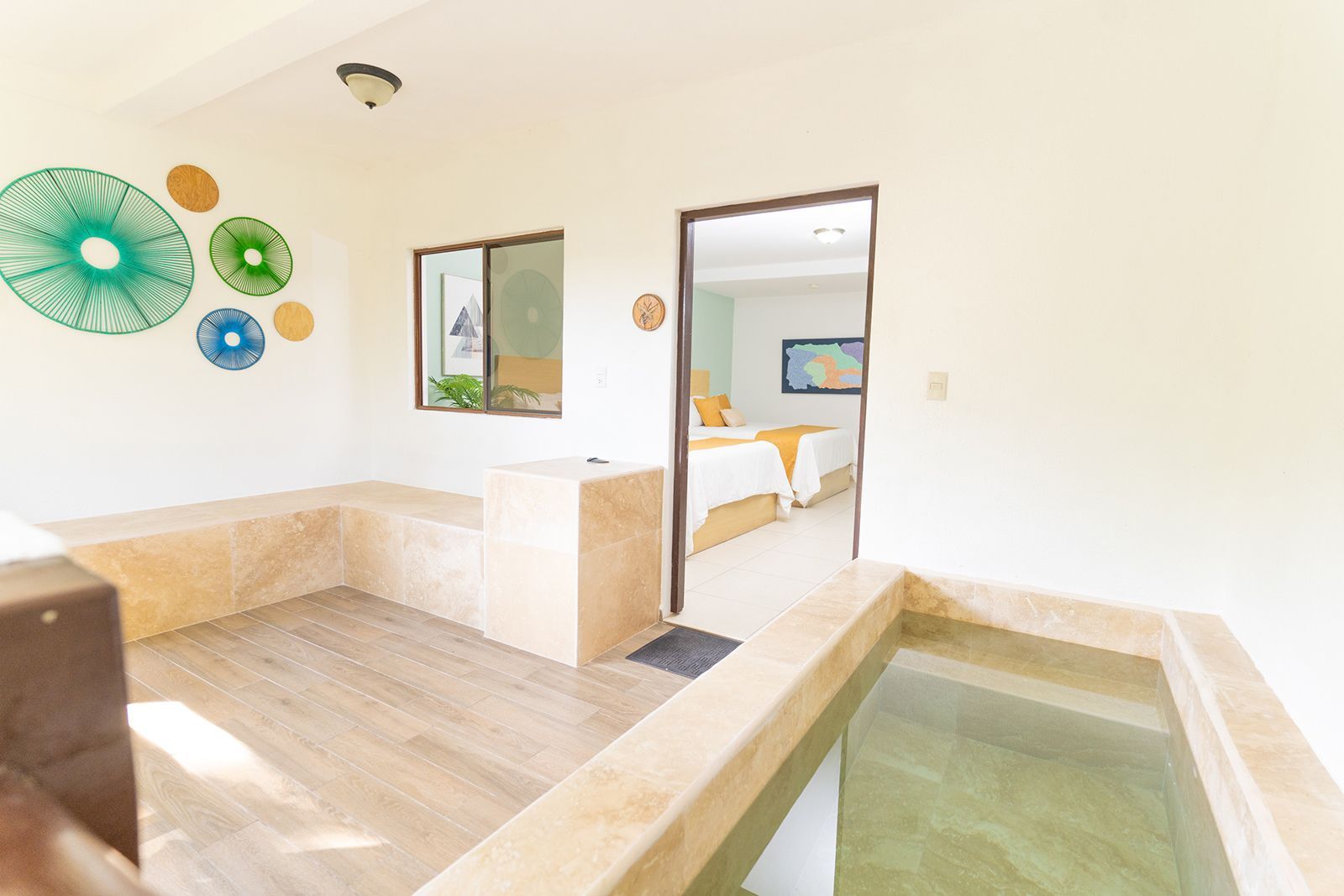 A sunlit spa room with a stone pool and a doorway revealing a bedroom with two beds, decorated with green wall art.