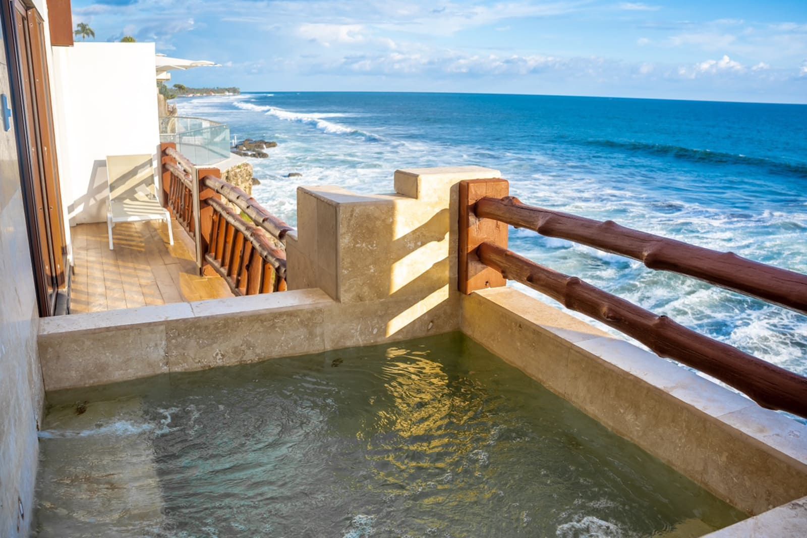 A small, rectangular stone pool on an outdoor balcony overlooking a blue ocean with waves crashing against the shore.