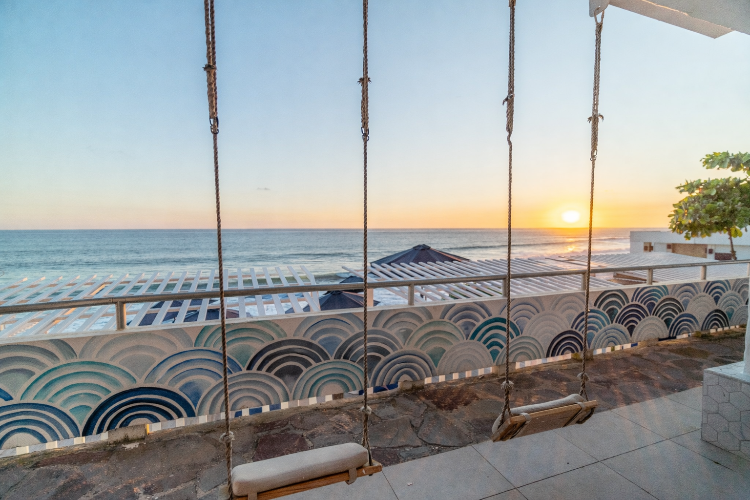 Two wooden swings hang in front of a sea-patterned wall, overlooking the ocean at sunset.