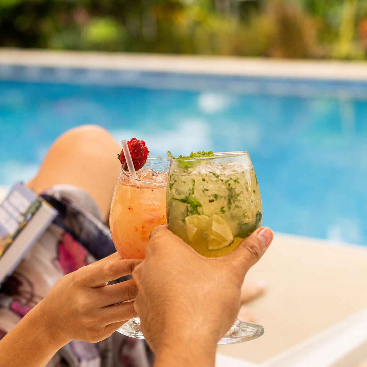 Two hands clinking tropical cocktails by a pool, with a blurred blue swimming pool in the background