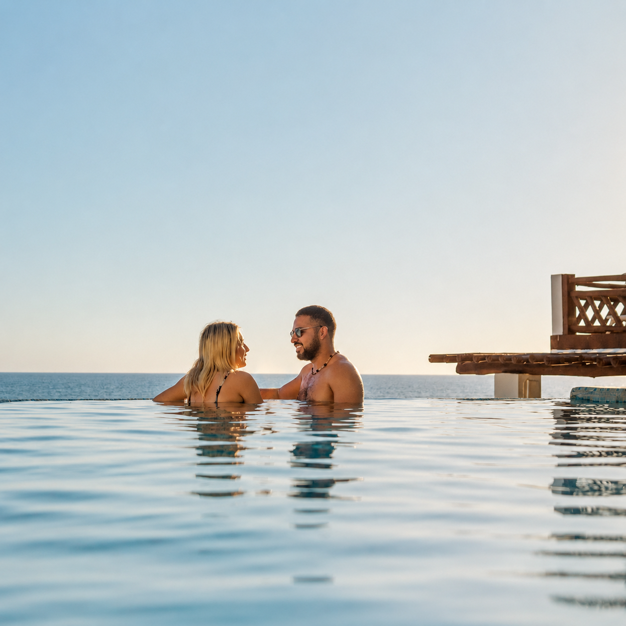 Two people chatting in an infinity pool beside a wooden deck at sunset