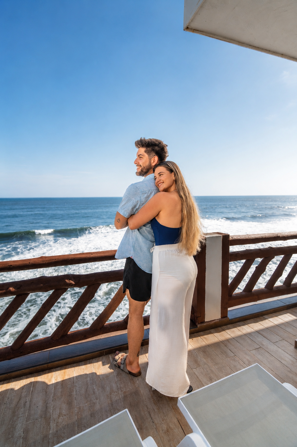 Couple embracing on a wooden deck overlooking the ocean under a clear blue sky.