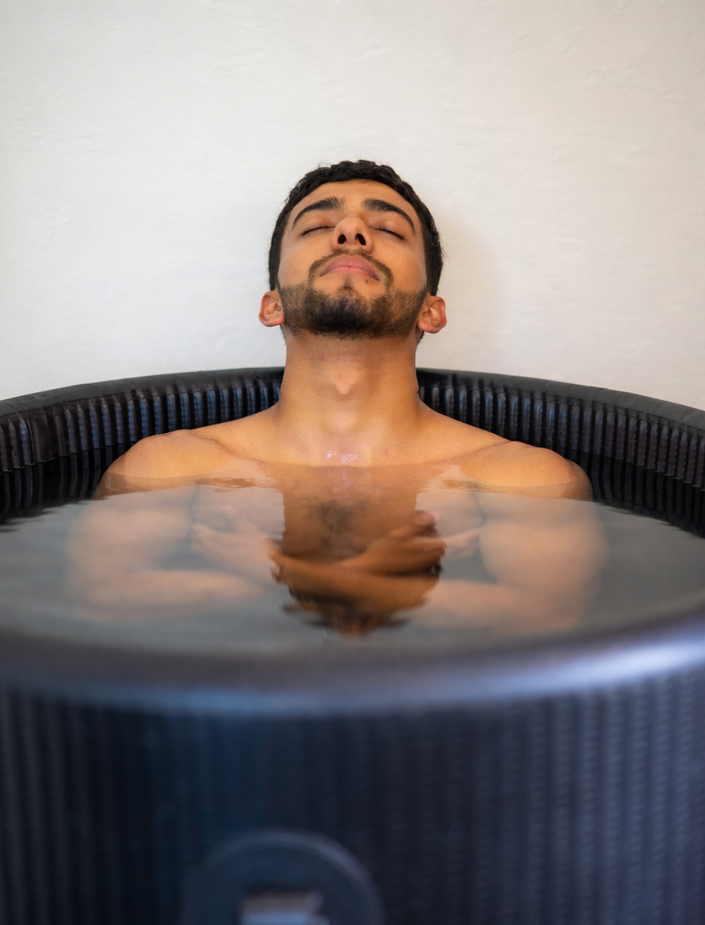 Person relaxing in a round cold plunge tub with head tilted back against a plain wall