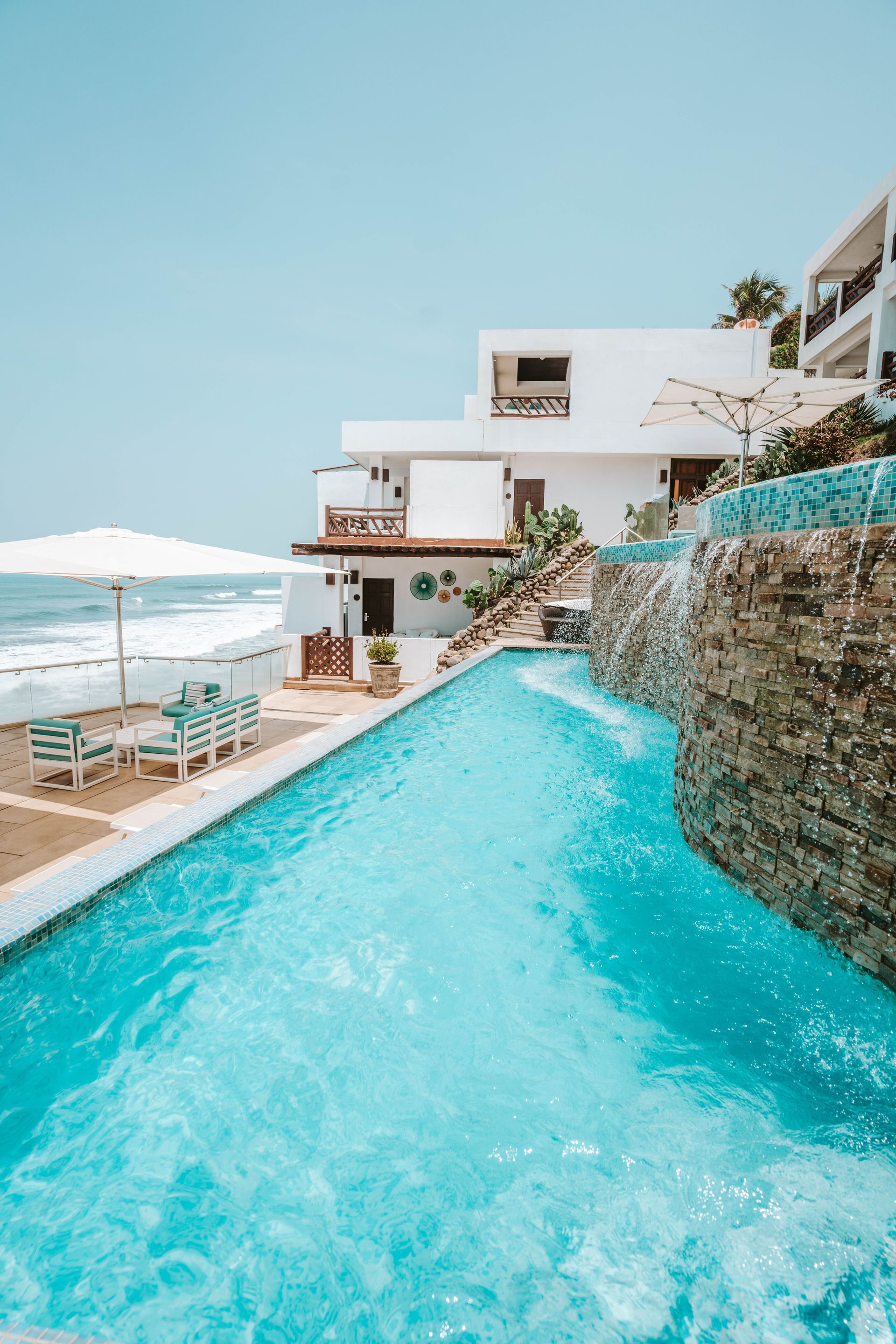A bright blue infinity pool overlooking the ocean, next to a white multi-level building with an outdoor lounge area.