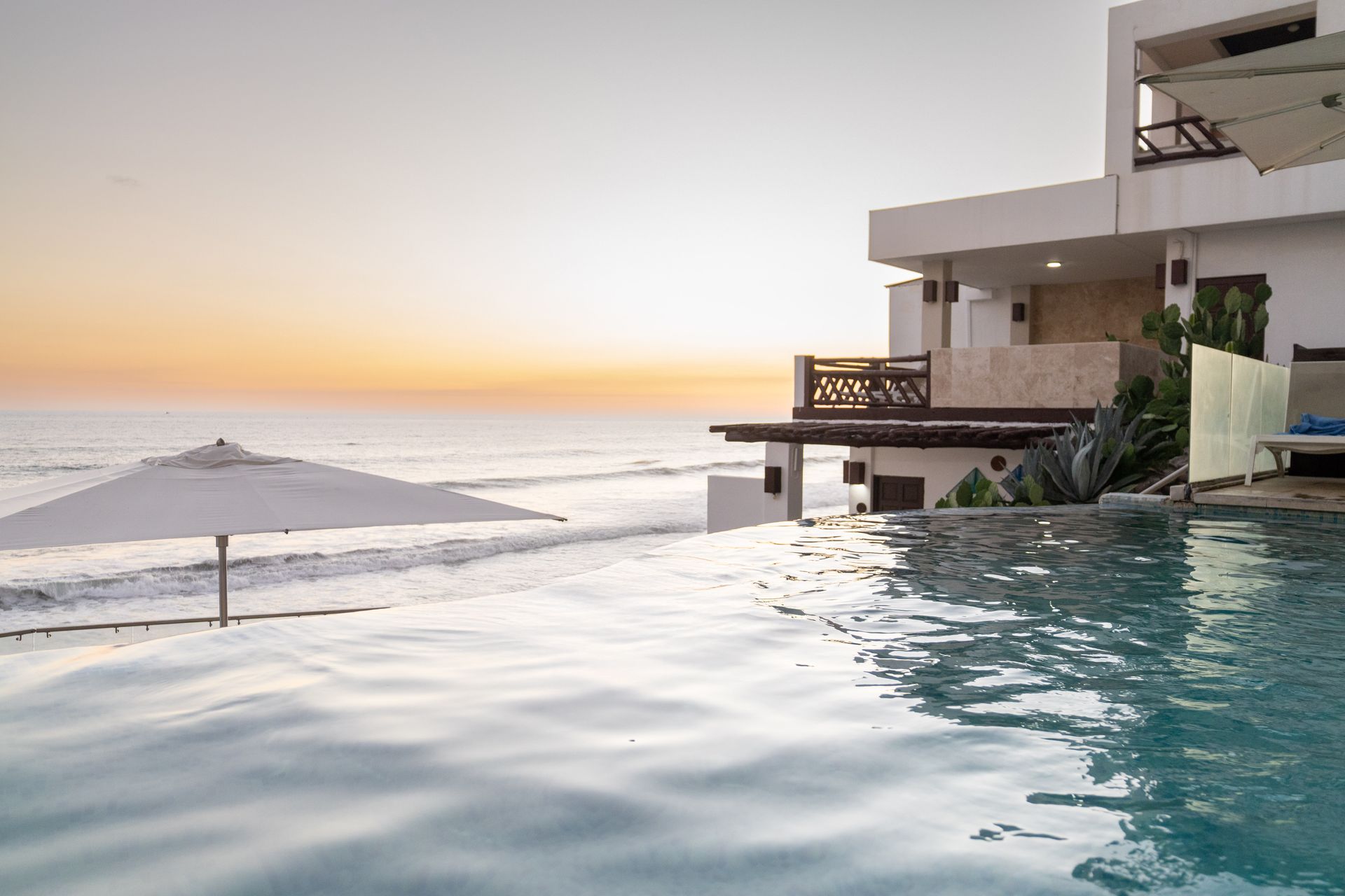 An infinity pool overlooking the ocean at sunset with a modern multi-story building to the right.