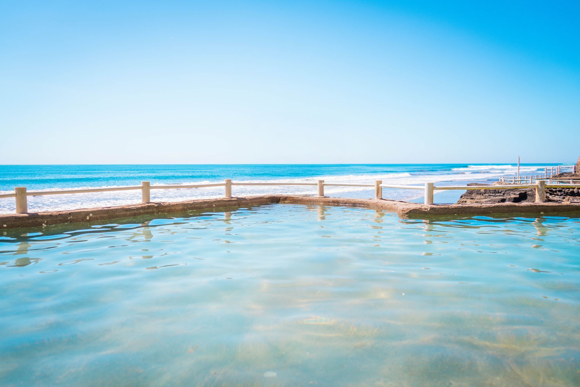 A calm, turquoise infinity-style swimming pool overlooking the ocean under a clear blue sky.