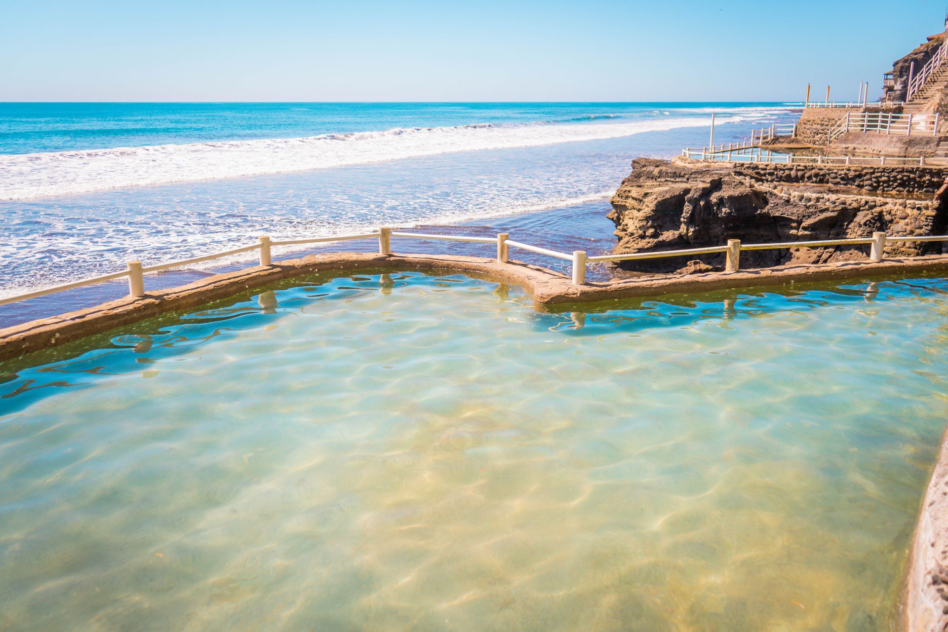A swimming pool with calm, light-blue water bordered by a low wall and railing, situated next to a rocky cliff and ocean.