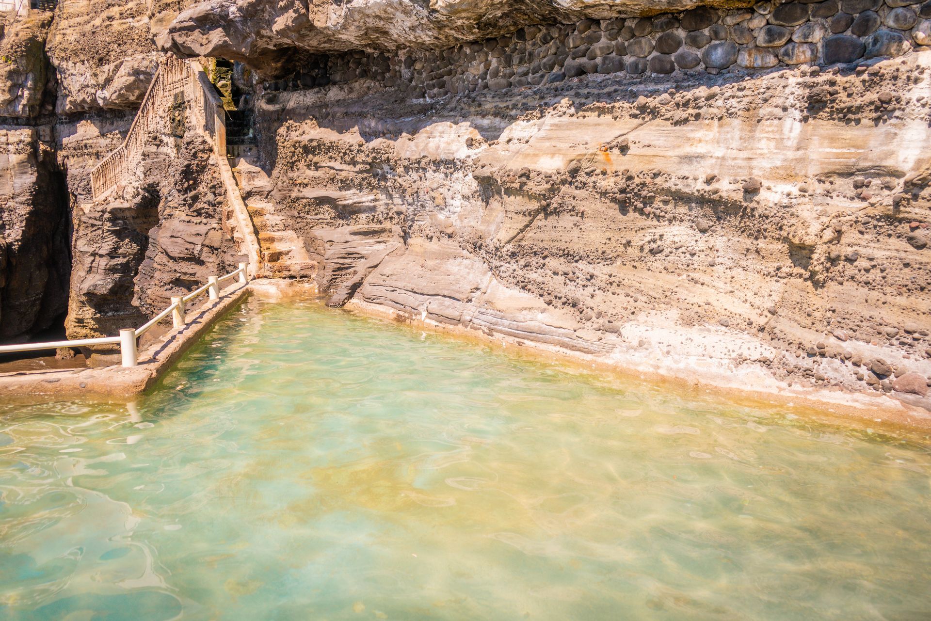 A calm, light-green swimming pool built into a natural, layered rocky cliffside with a stone staircase leading up.