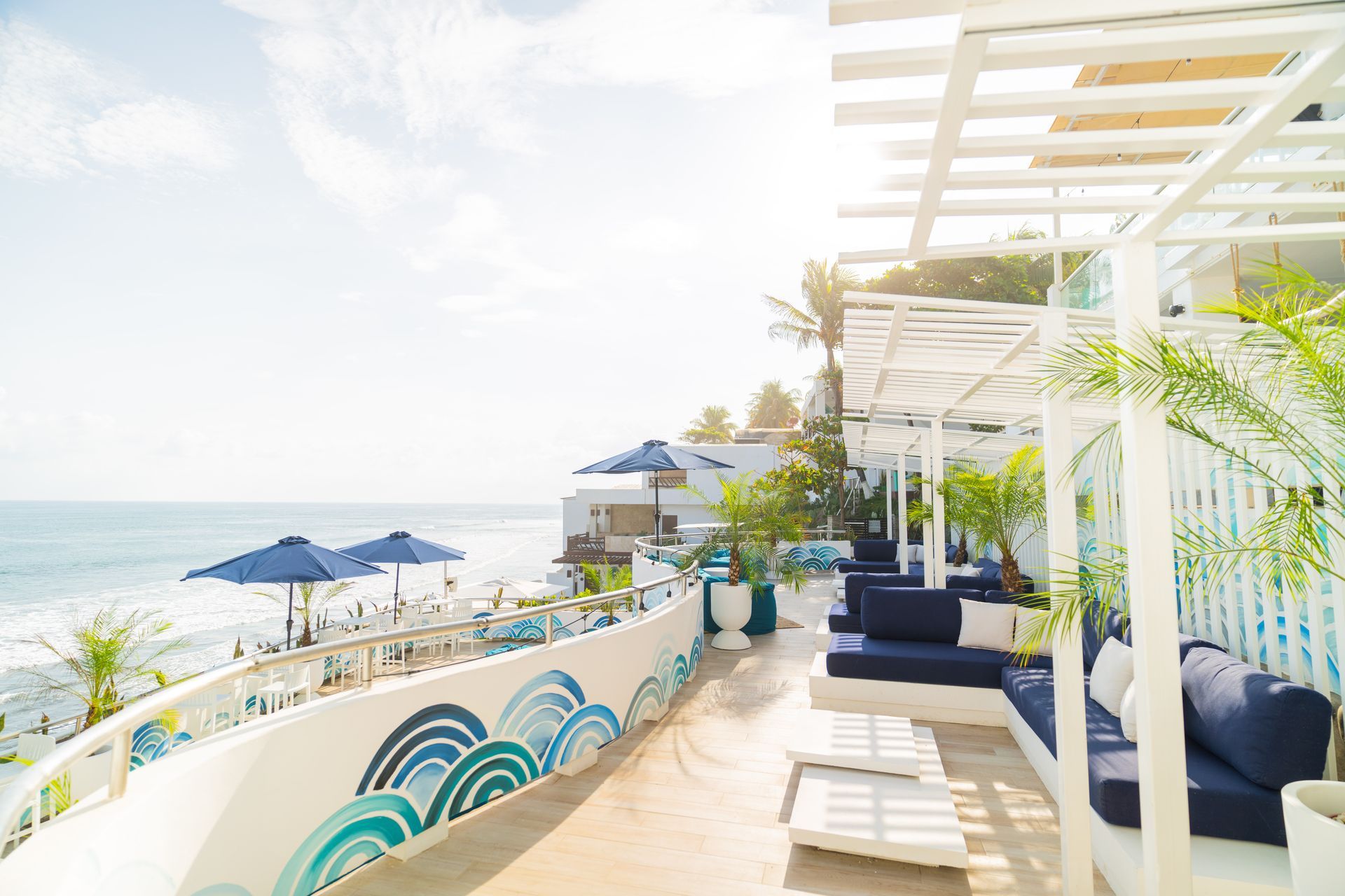 An outdoor seaside terrace featuring white pergolas, blue cushioned lounge seating, and a decorative wave-patterned wall.