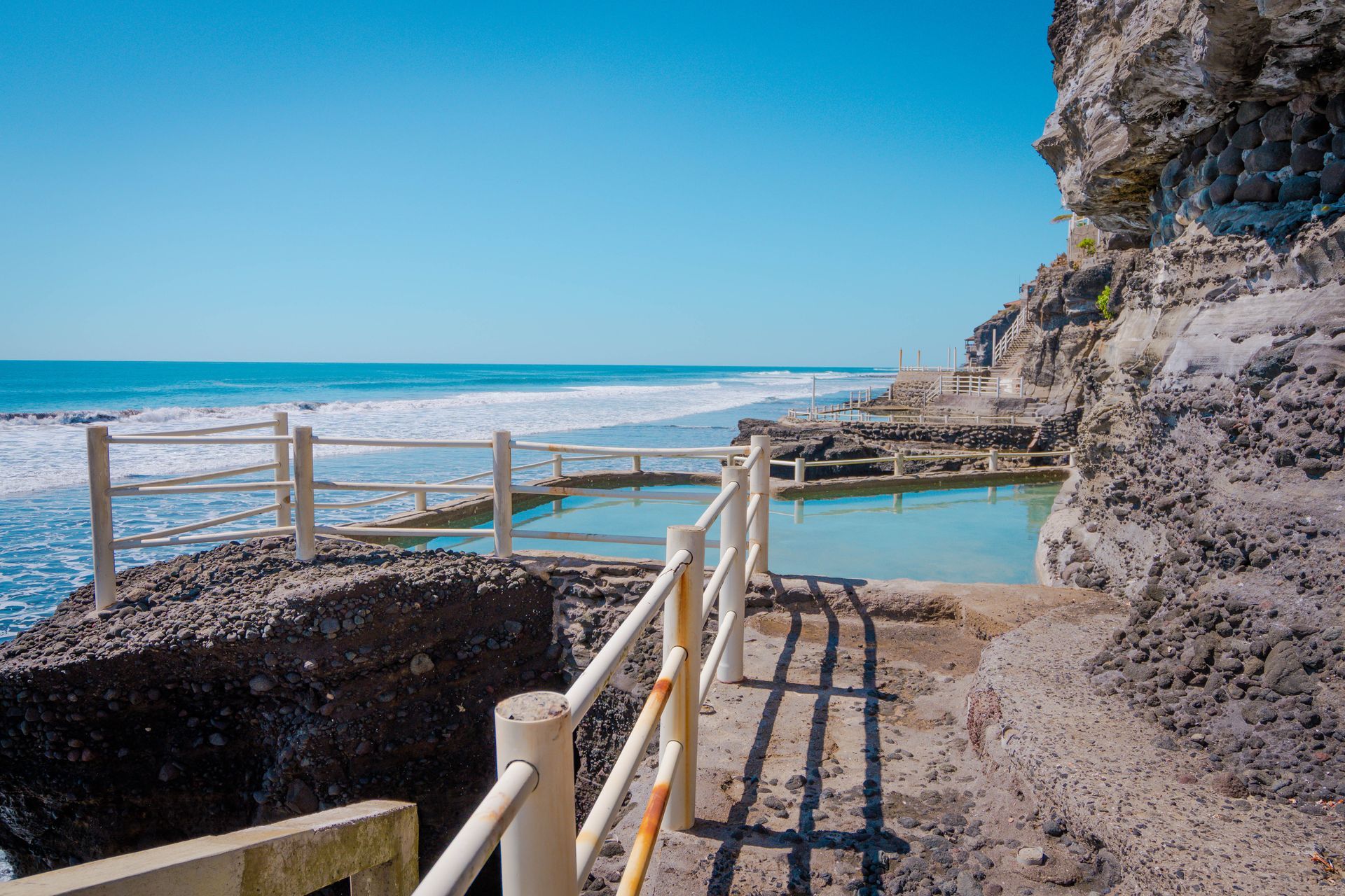 A natural tide pool filled with turquoise water, situated on a rocky cliffside next to the ocean under a clear blue sky.