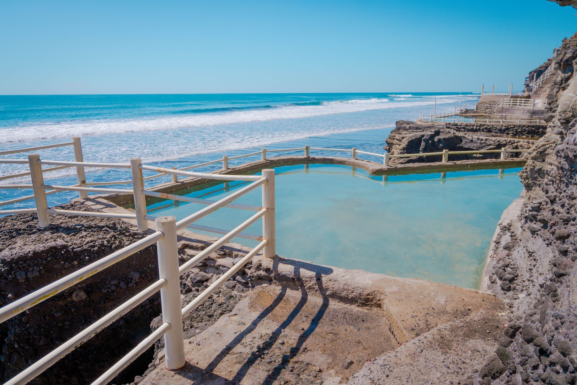 A turquoise natural tidal pool carved into rocky coastal cliffs beside a bright blue ocean under a clear sky.
