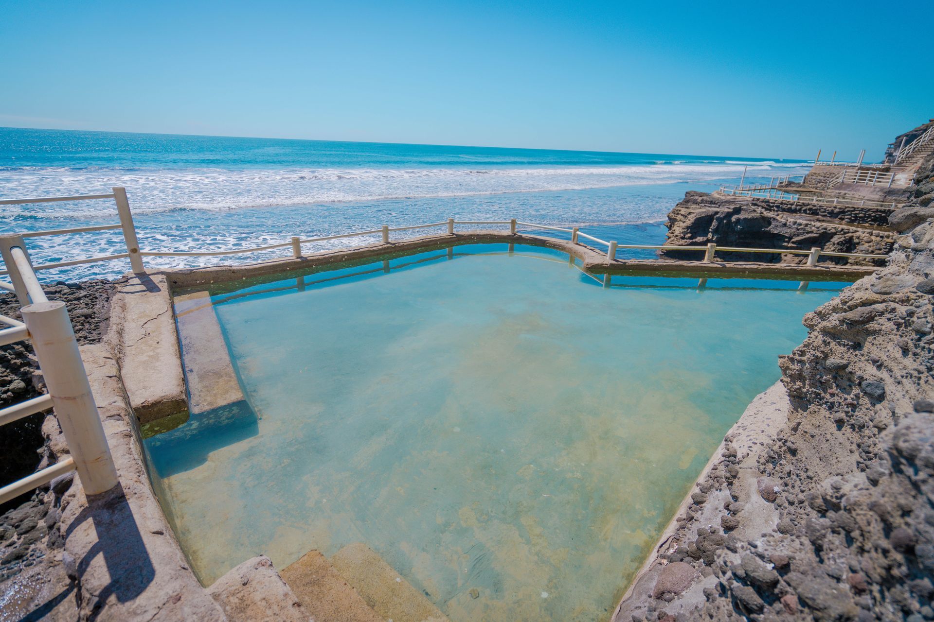 A concrete pool filled with turquoise water sits along a rocky coastline next to the ocean under a clear blue sky.