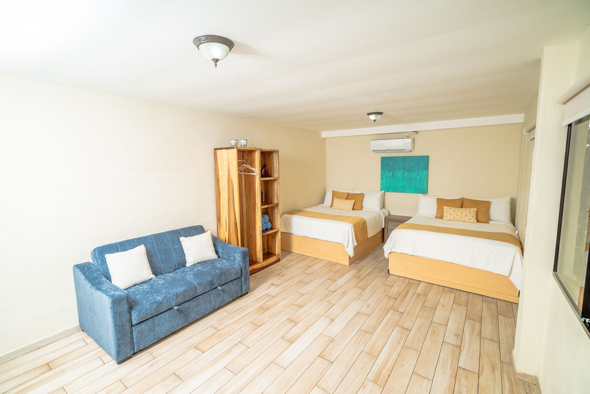 Bright bedroom with two single beds, blue sofa, wooden floor, and a window with teal curtain.