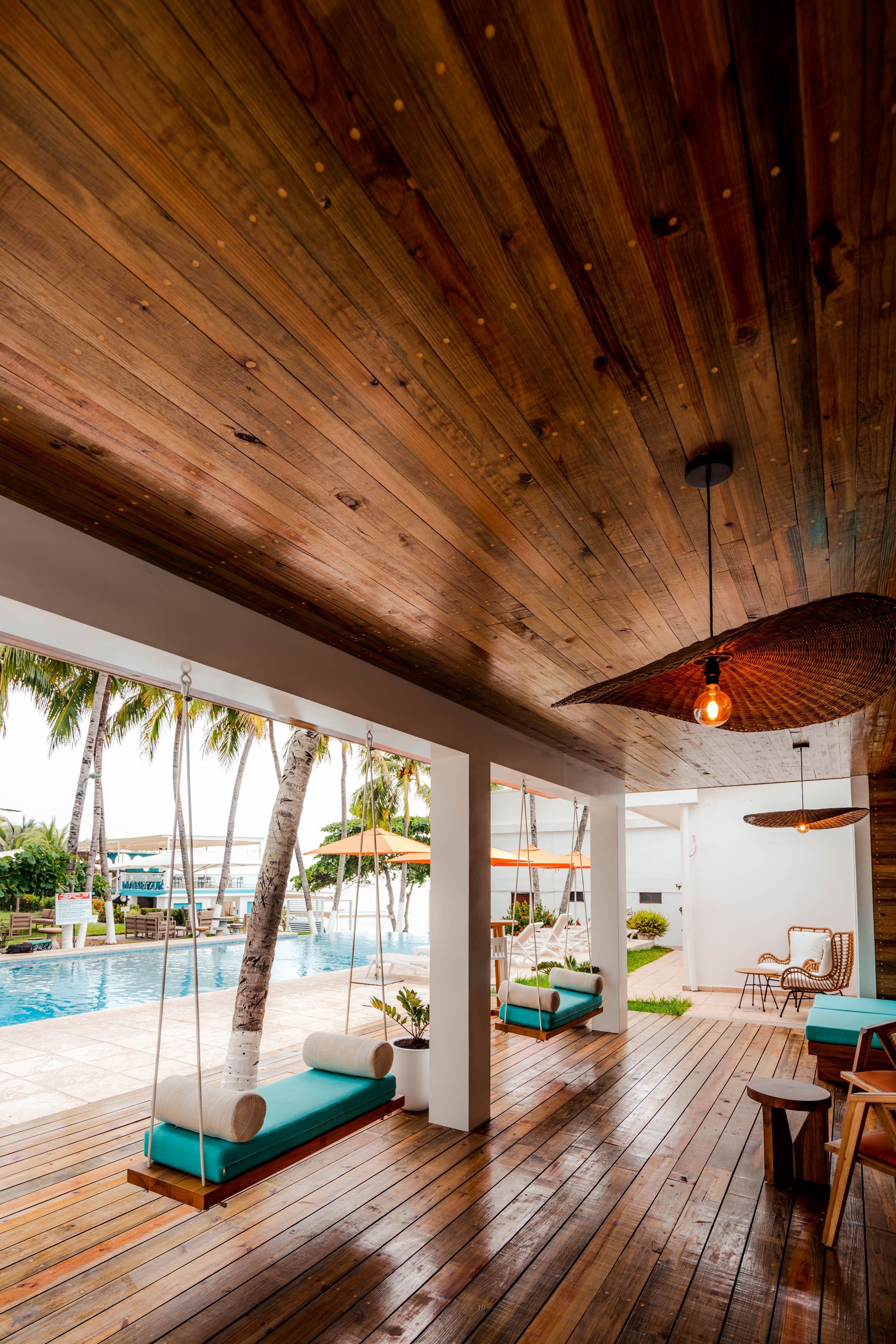 A wooden deck with hanging swing beds overlooking a swimming pool and tropical trees on a bright, sunny day.