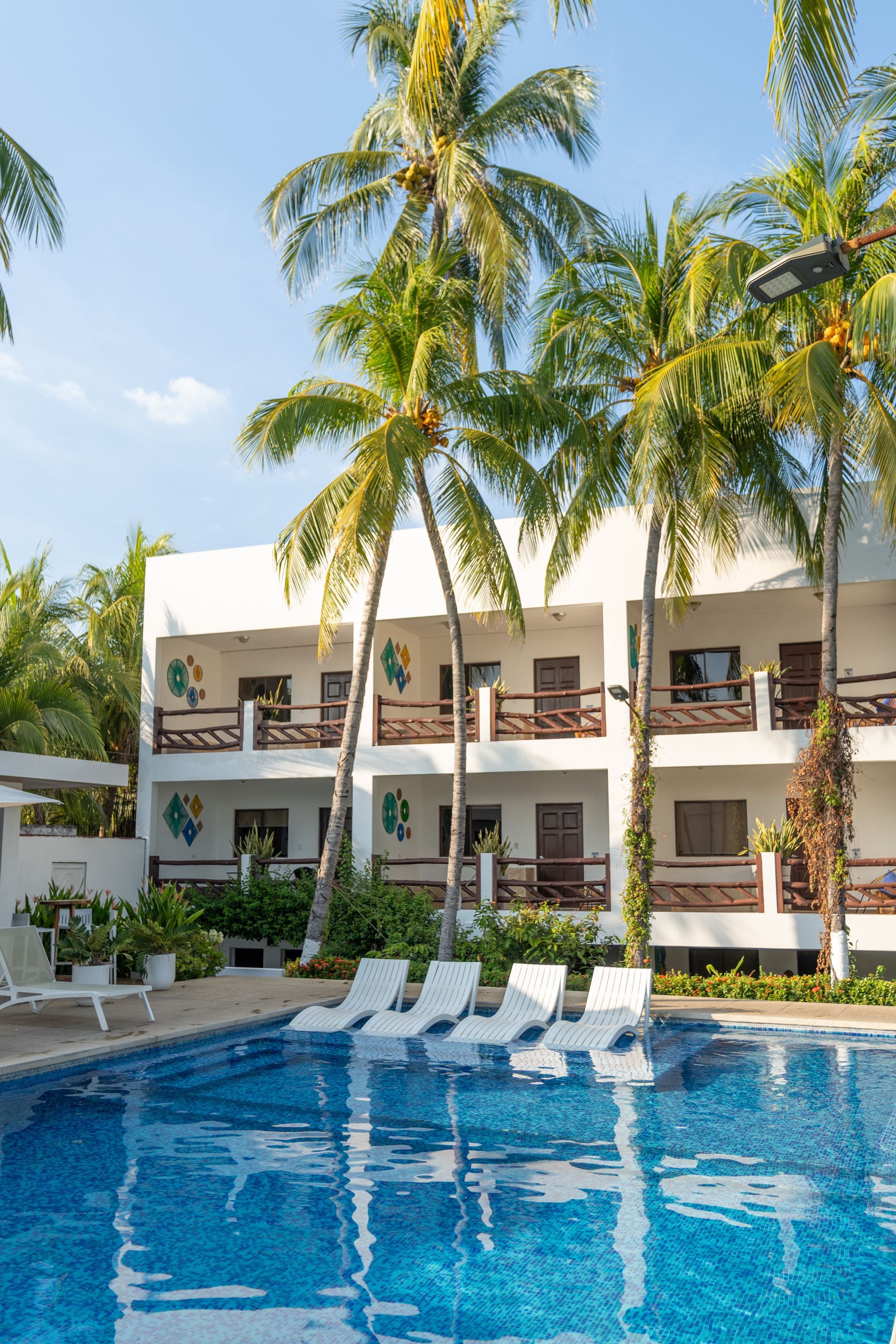 A swimming pool with lounge chairs sits in front of a white two-story hotel surrounded by tall palm trees under a blue sky.