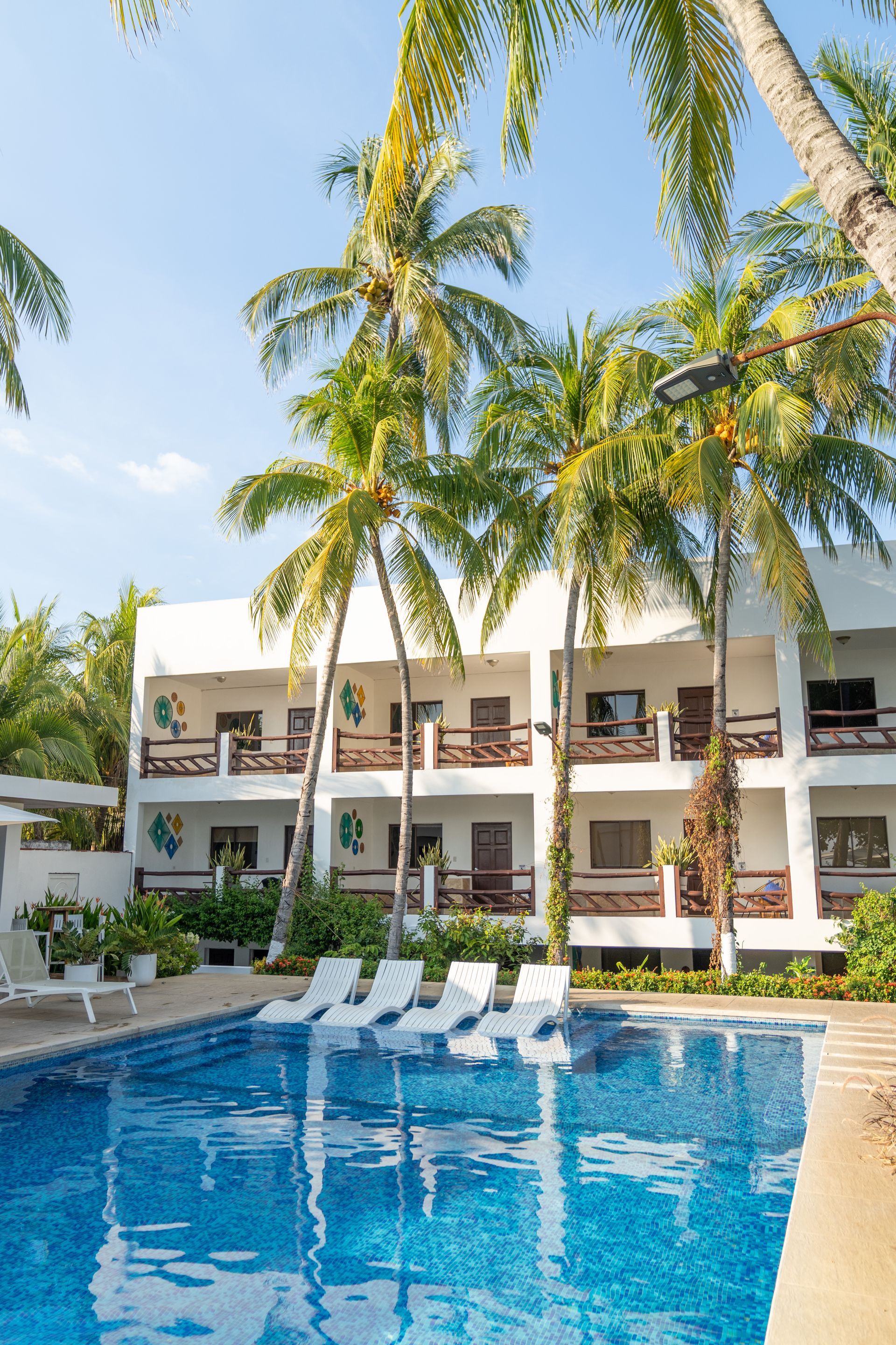 A resort building with white balconies and palm trees situated behind a blue swimming pool with lounge chairs.