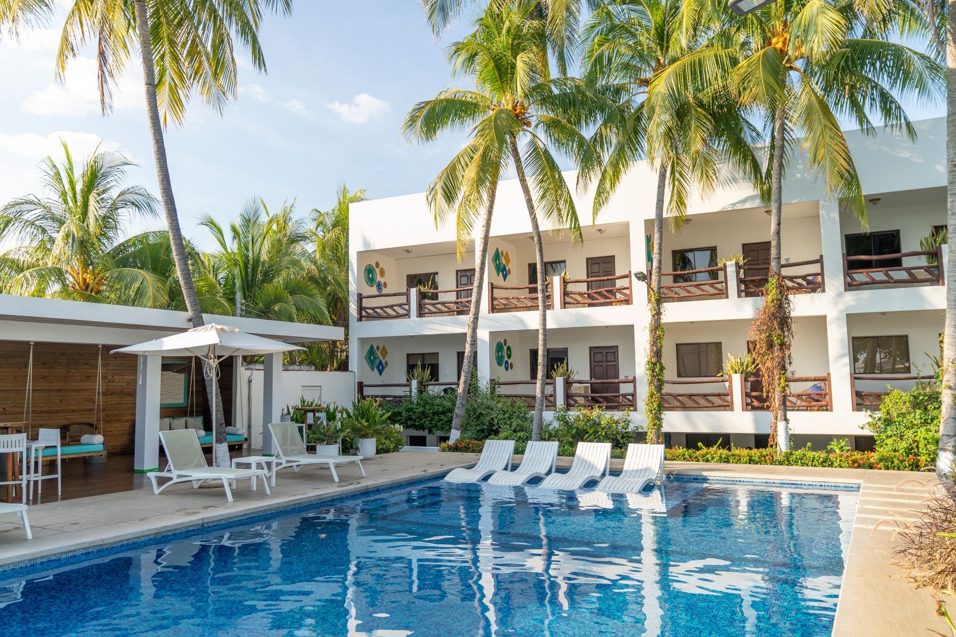A swimming pool surrounded by lounge chairs, palm trees, and a three-story white hotel building.