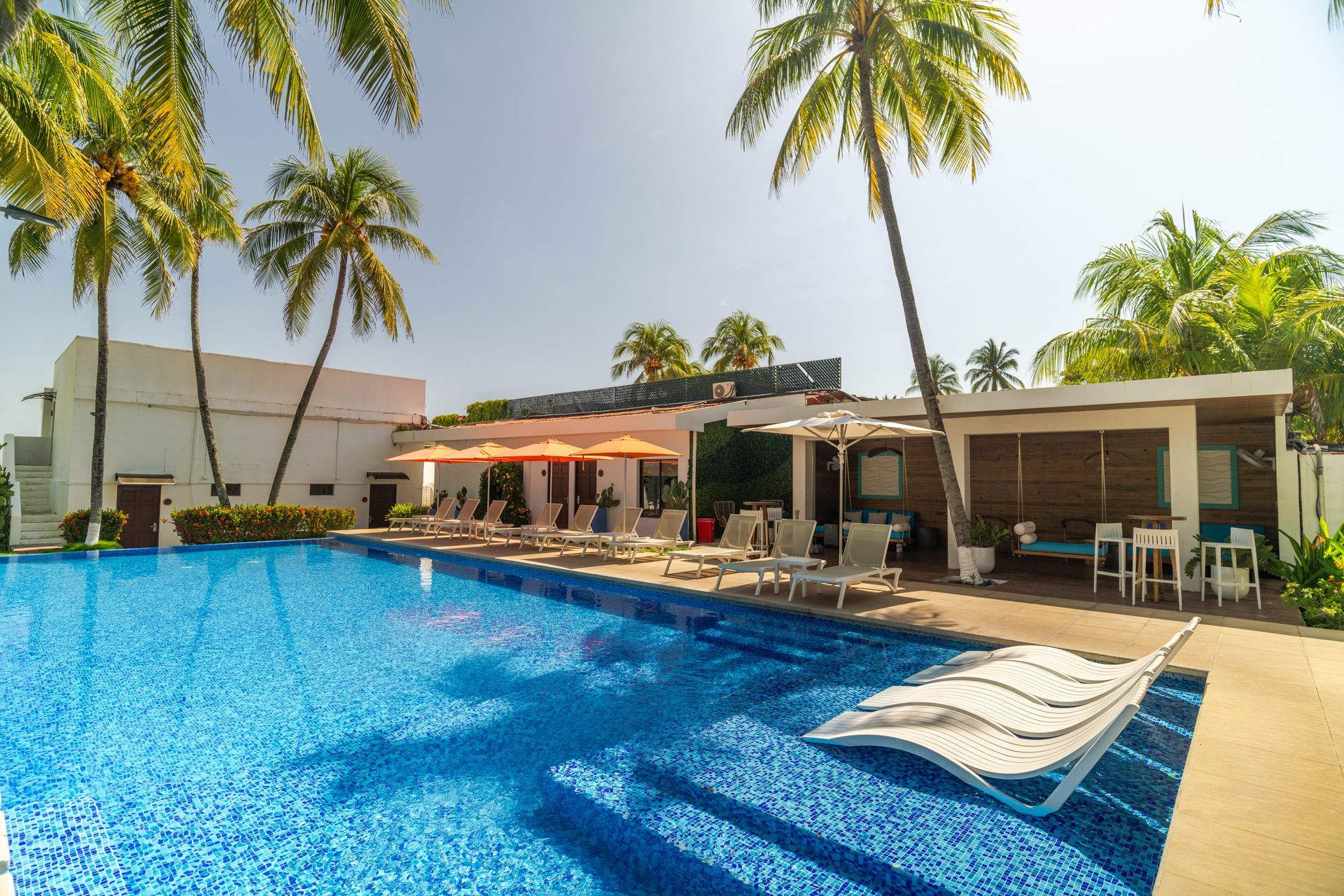 A sunny, tropical resort swimming pool with bright blue water, lounge chairs, palm trees, and an open-air dining area.