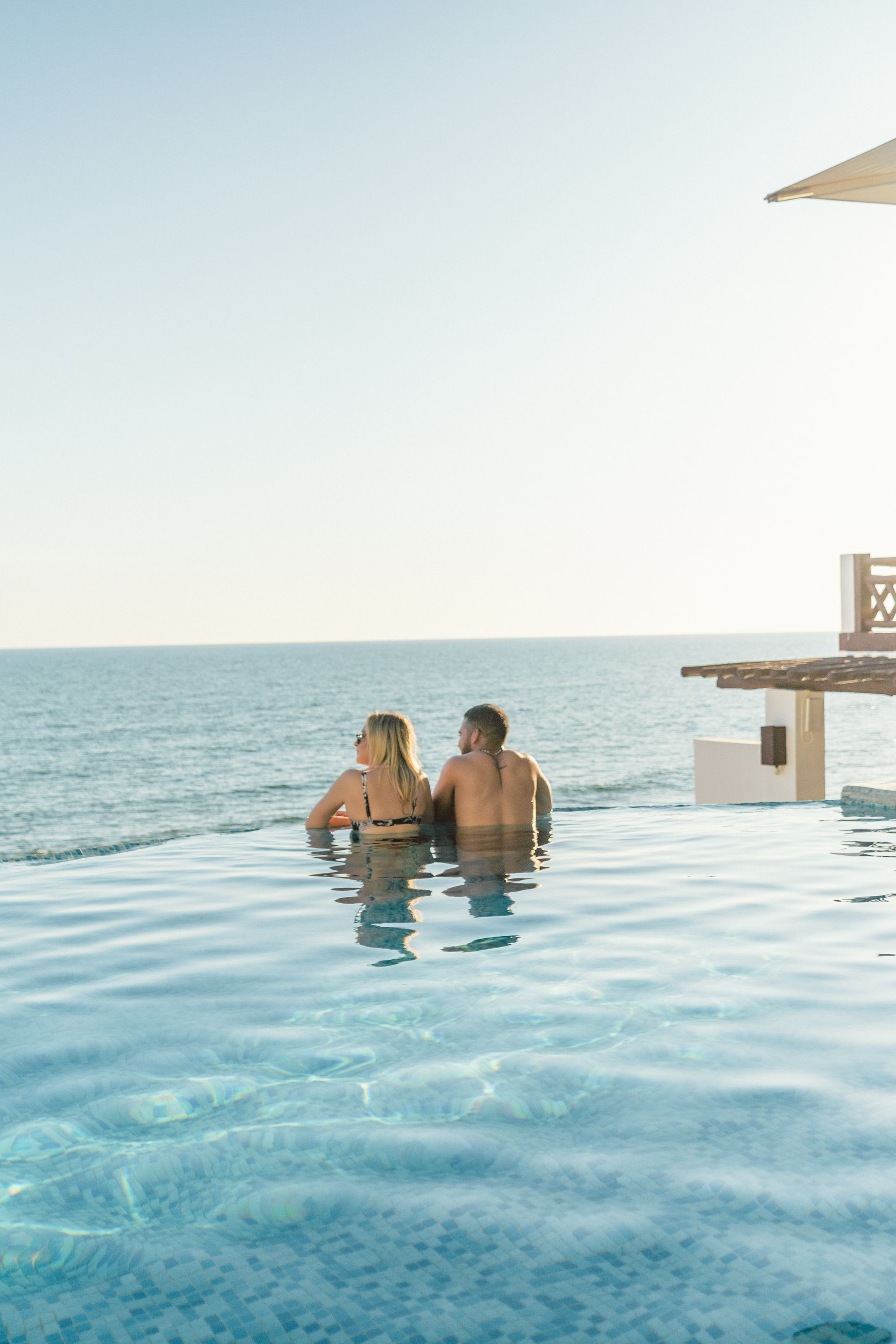 Two people relax in an infinity pool overlooking a calm ocean at sunset.