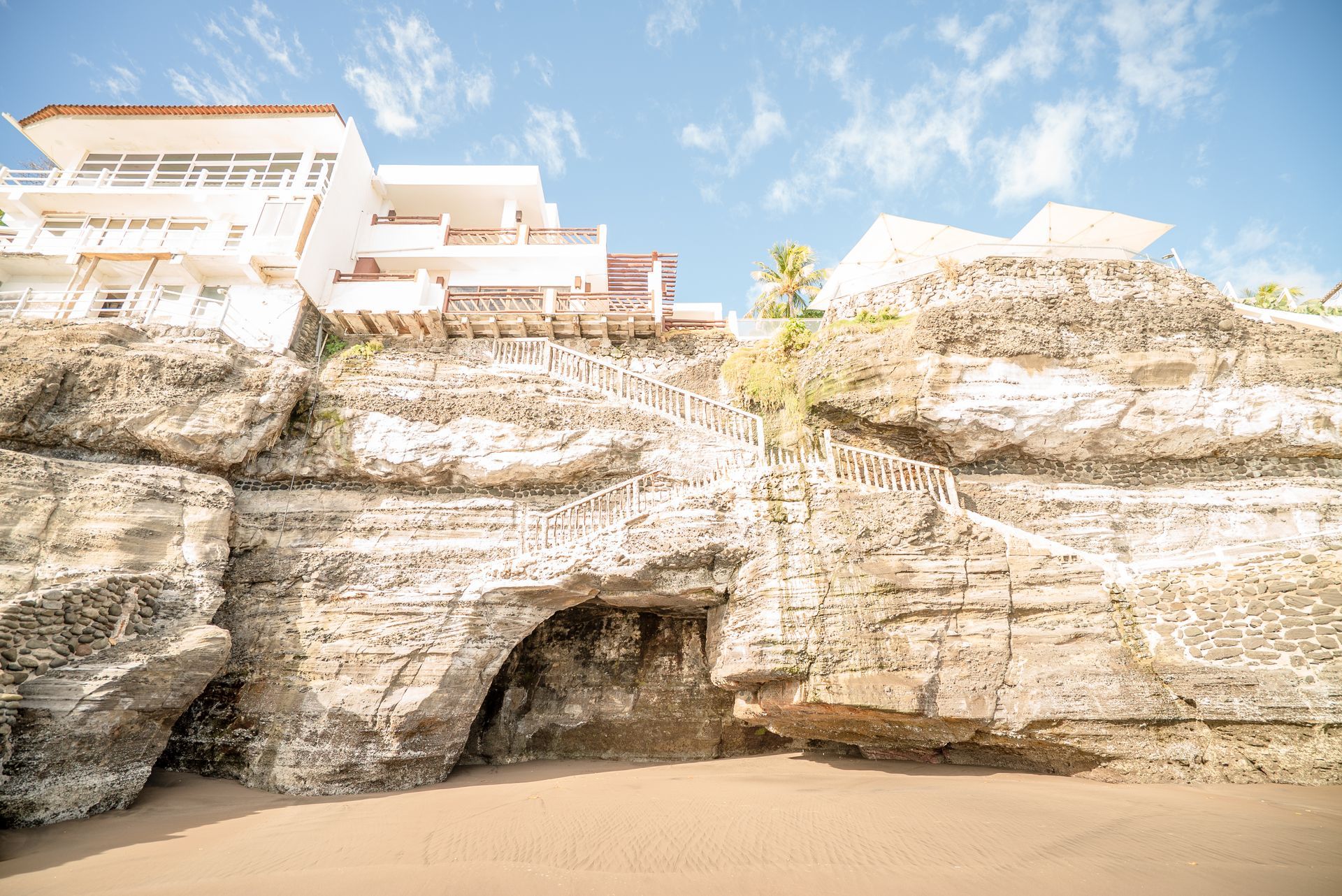 A white building sits atop a rugged, textured cliff overlooking a sandy beach with a small natural archway.