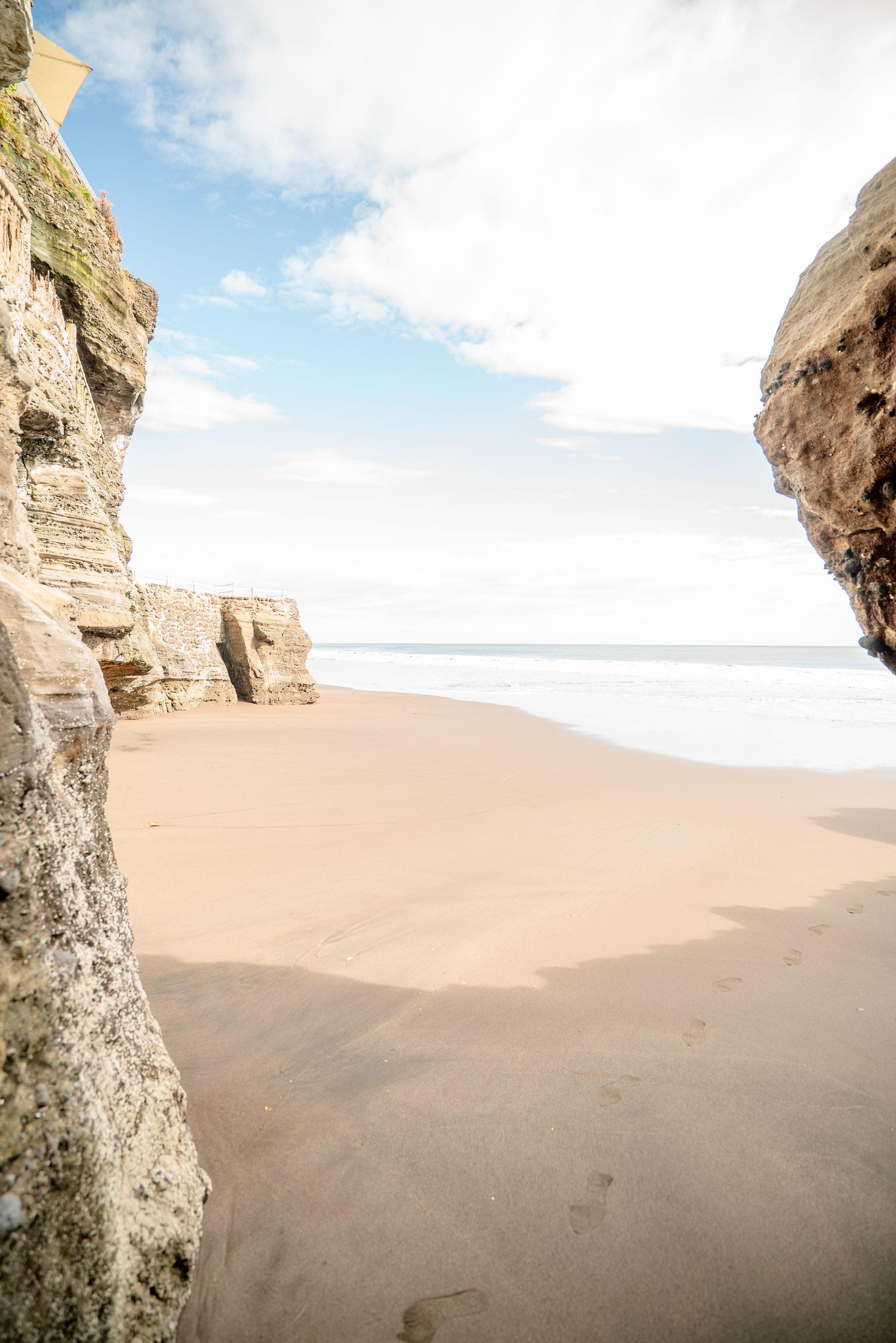 A sandy beach viewed through a narrow opening between two large, weathered rock formations under a bright, cloudy sky.