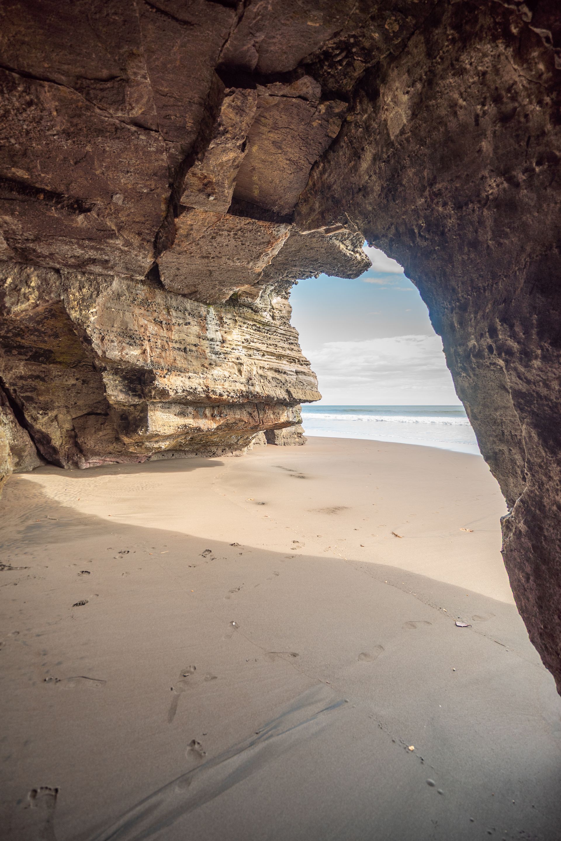 A sandy beach framed by a rocky sea cave, looking out toward the ocean under a cloudy sky.