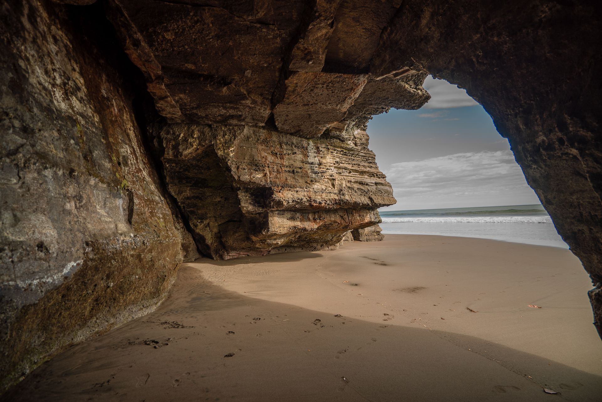 A sandy beach viewed from inside a rocky cave, with a view of the distant coastline under a cloudy sky.
