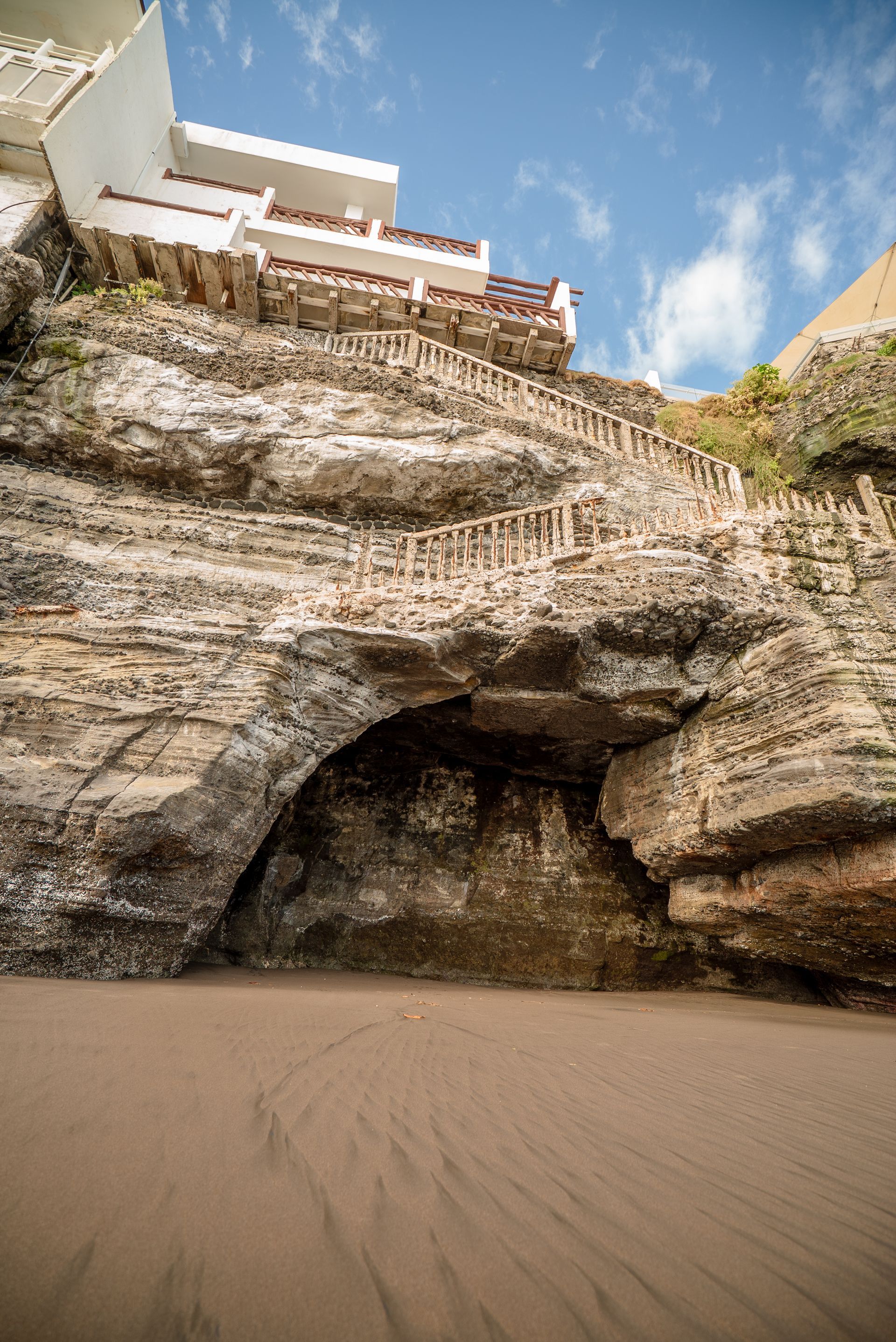 A sandy beach leads to a dark sea cave carved into a tall, rugged limestone cliff topped by a white building.
