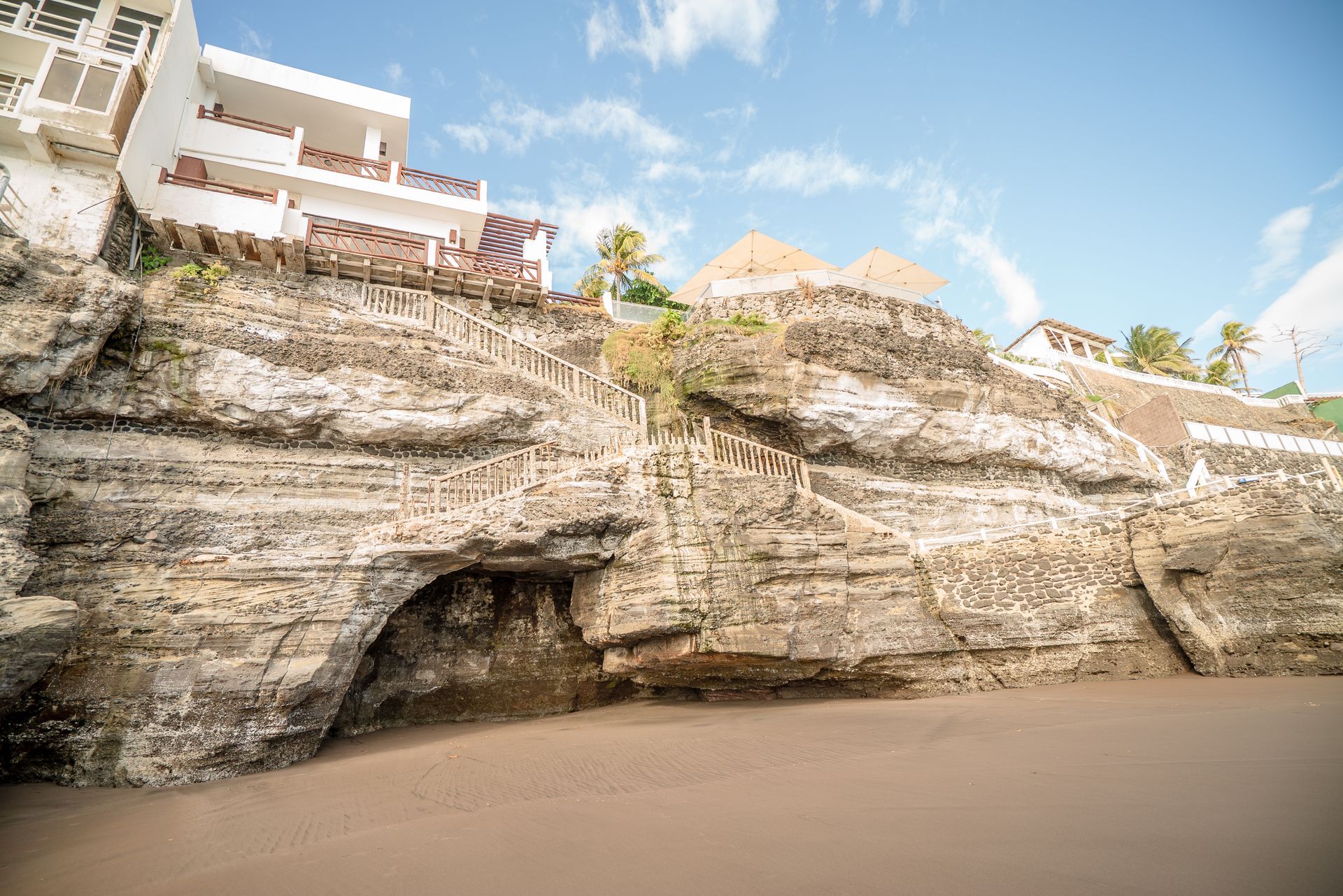 A rocky limestone cliff with a small cave opening, rising above a flat sandy beach beneath a bright blue sky.