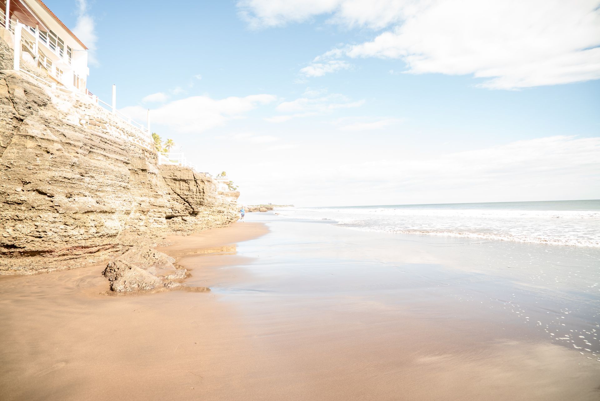 A sandy beach with rocky cliffs on the left, leading to a calm sea under a bright, sunny sky.