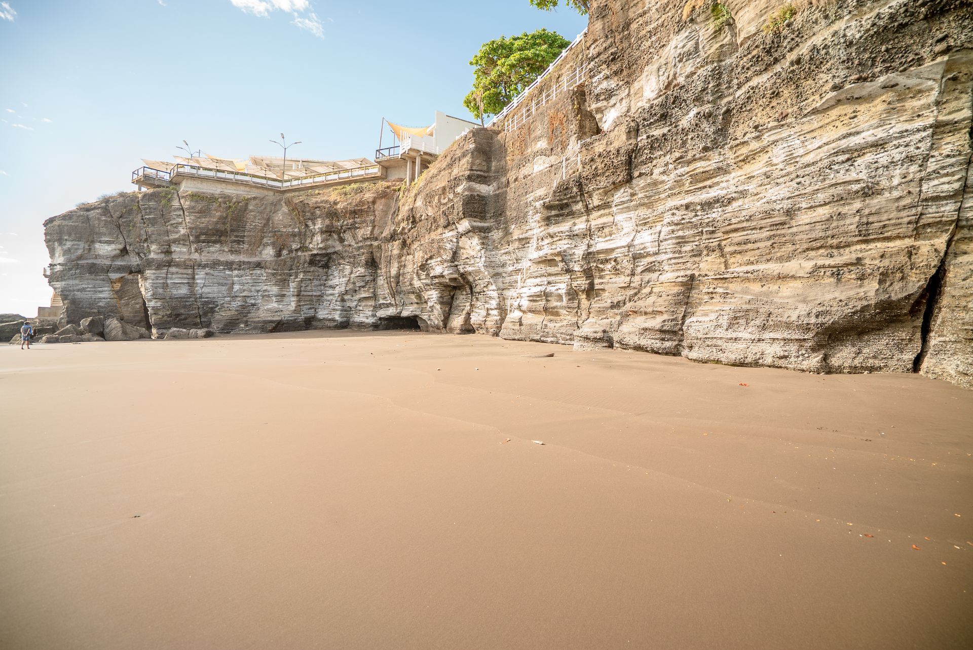 A wide, flat sandy beach beneath a tall, jagged cliff face under a clear blue sky.