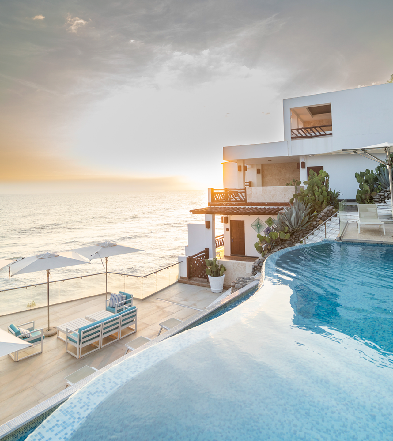 Infinity pool overlooking the ocean at sunset, featuring a terrace with lounge furniture and a white modern villa.