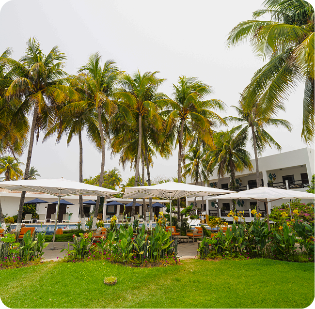 A white resort building with palm trees, outdoor umbrellas, and lush green landscaping surrounding a pool.