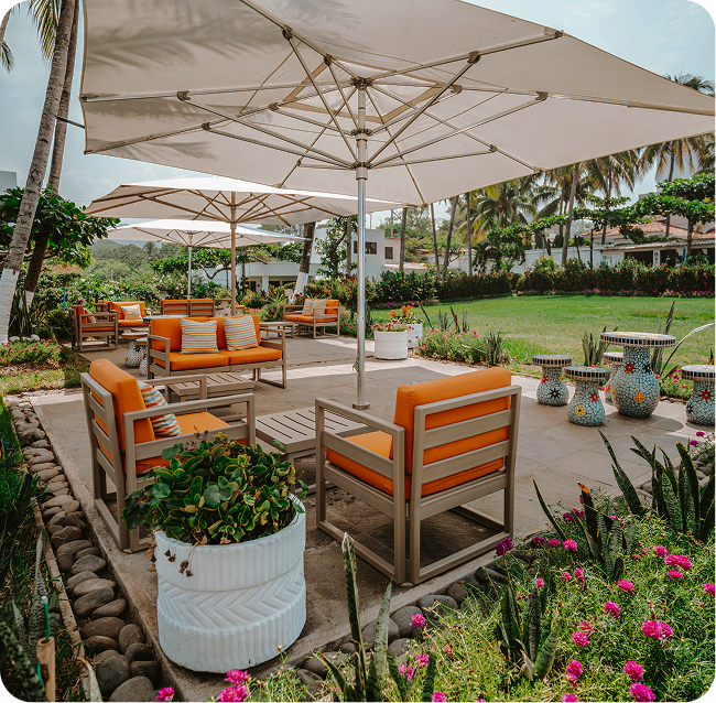 Outdoor patio with orange cushioned chairs under large white umbrellas, set on a stone deck surrounded by garden plants.