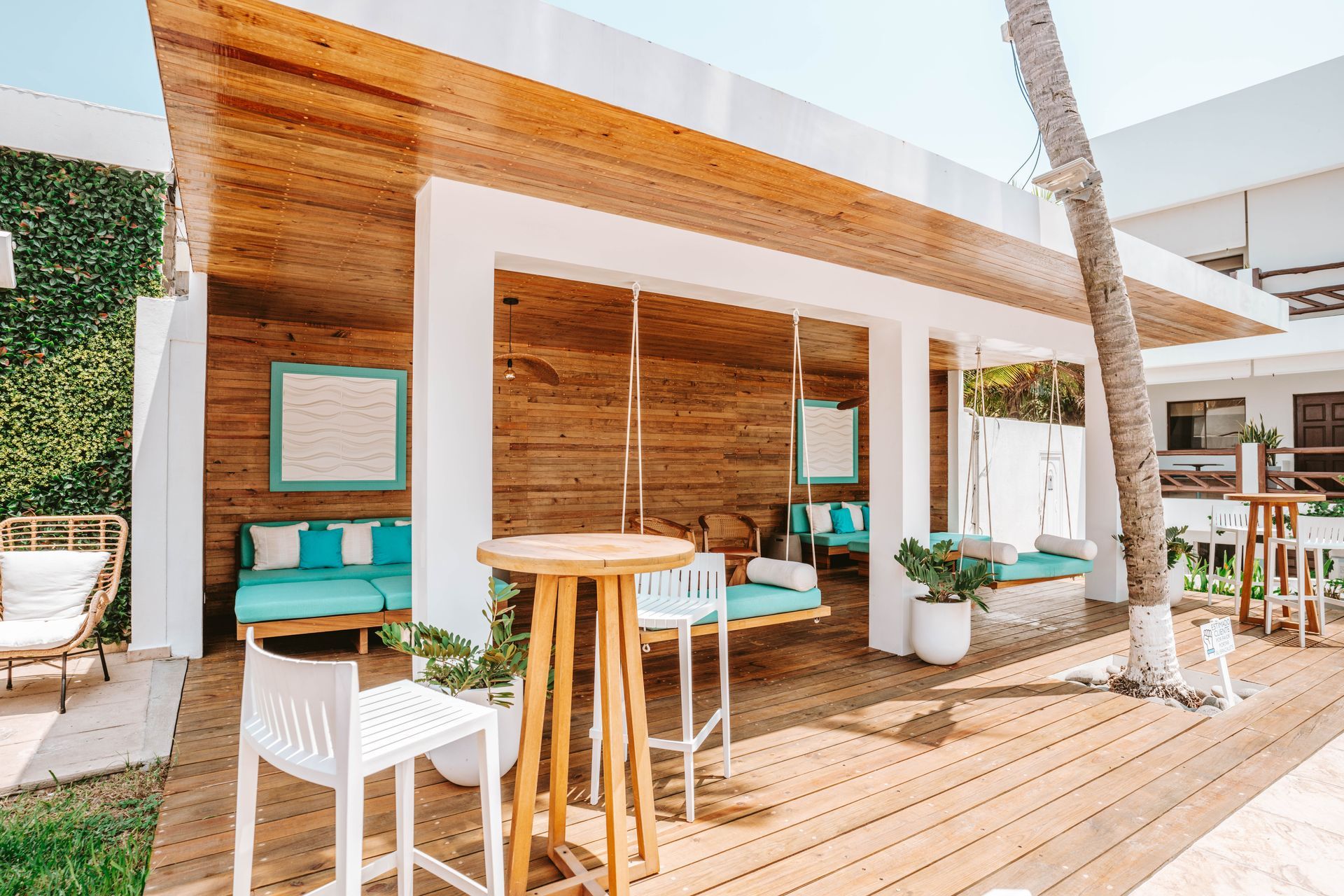 A covered outdoor patio with wooden walls and flooring, featuring swinging benches, tables, and white chairs.