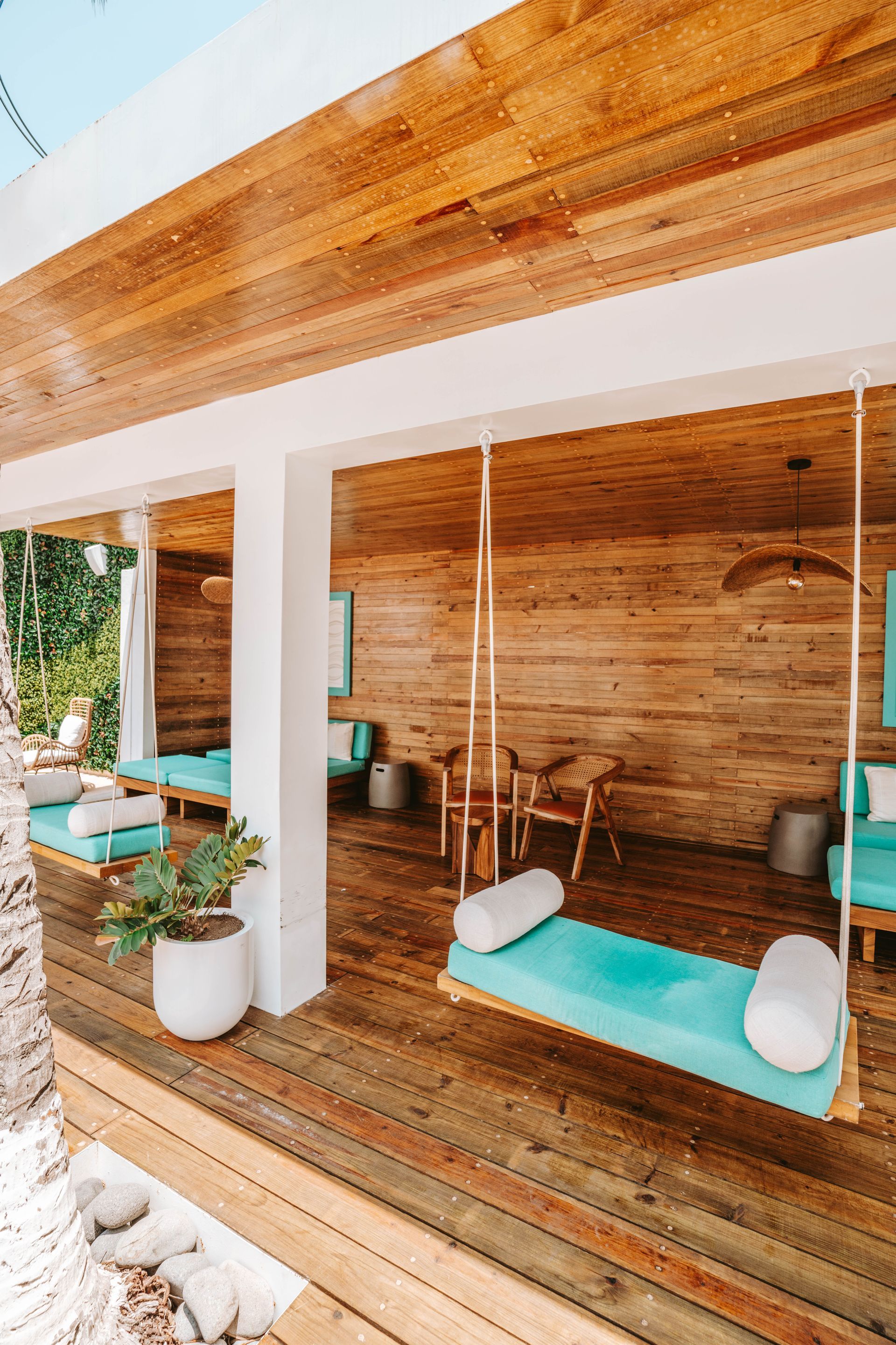 A wooden open-air patio features hanging turquoise daybeds and chairs, with a potted plant and tropical surroundings.