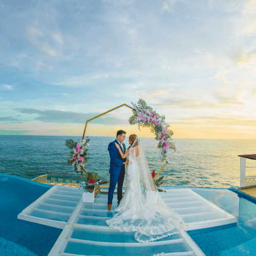 A couple stands on a platform over a pool at sunset, framed by a gold hexagonal wedding arch decorated with flowers.