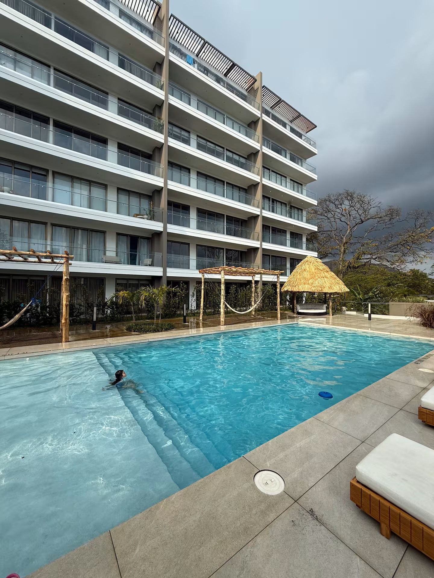 Modern hotel pool beside a multi-story balcony building under cloudy skies