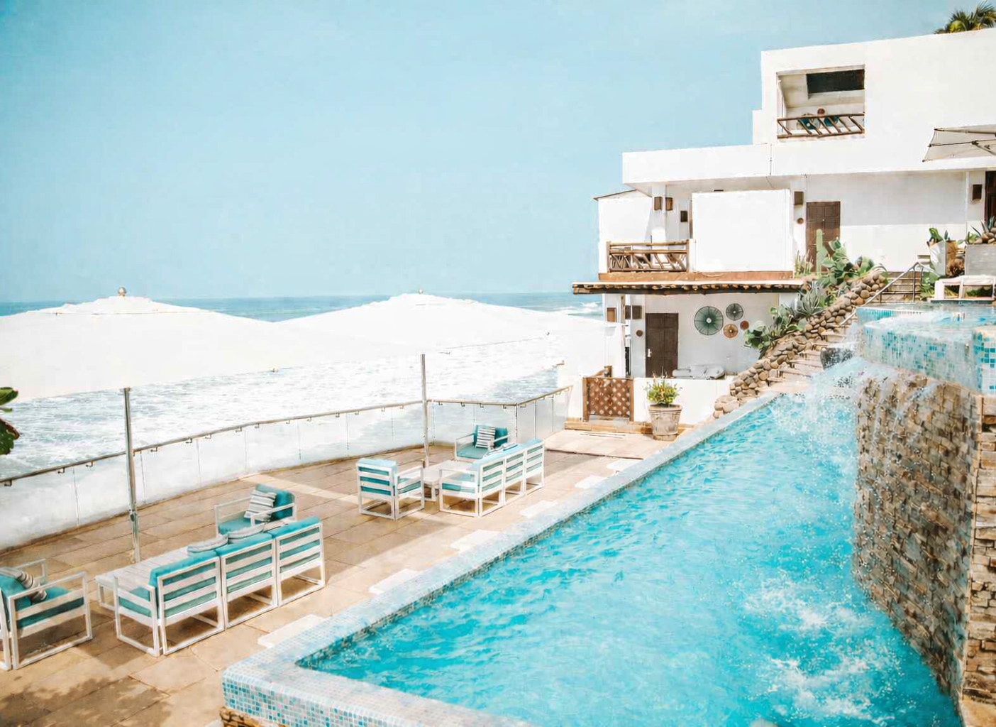 An infinity pool overlooking the ocean, with white lounge chairs under umbrellas and a multi-level white resort building.