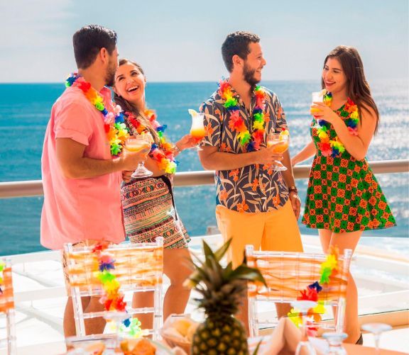 Four people in tropical attire and leis laugh together while holding drinks on an oceanfront deck.