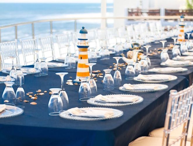 A long table set for a coastal-themed event with blue tablecloths, white plates, glasses, and lighthouse centerpieces.