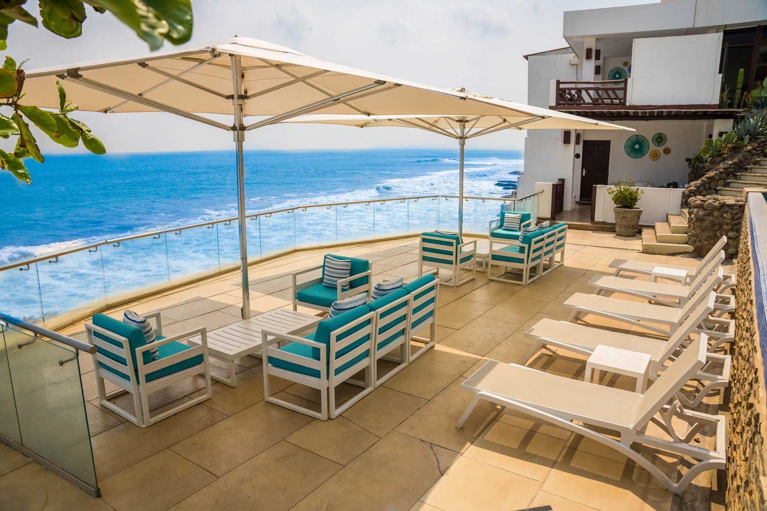 A bright patio overlooking the turquoise ocean with white lounge chairs, sofas, and large umbrellas.