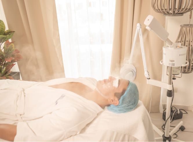 A person lying on a spa bed receives a facial steam treatment from a professional machine in a brightly lit room.