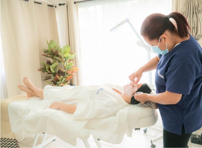 A professional in scrubs and a face mask performs a facial treatment on a client resting on a treatment table.