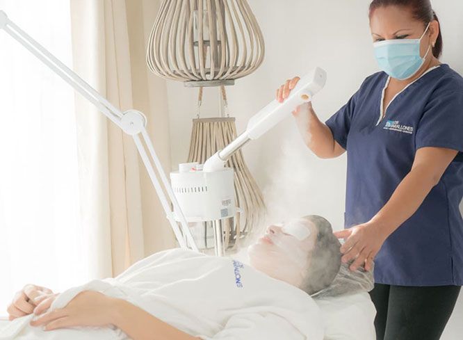 An esthetician in a blue uniform and face mask uses a facial steamer on a client lying on a treatment table.